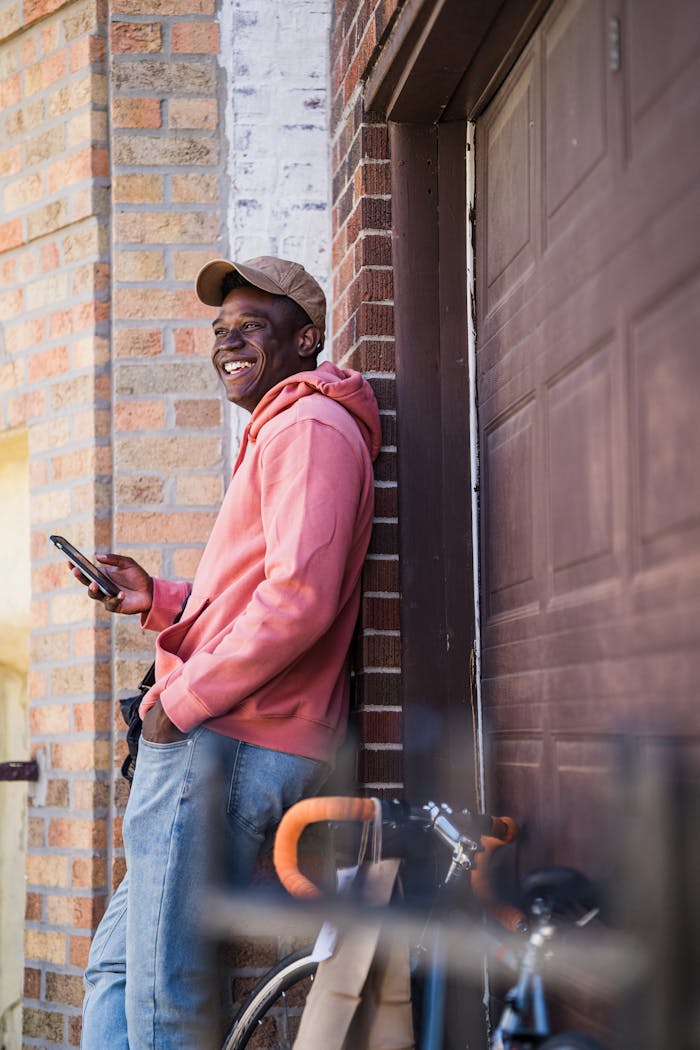 services-02 Man smiles while holding phone, leaning on brick wall with bicycle nearby.