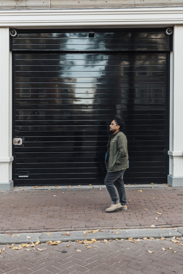about-us-01 Casually dressed man walks past black garage door in urban setting.